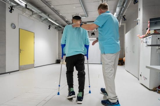 A rehab patient walks with crutches. His physiotherapist is next to him. He has his hand on the patient's back.
